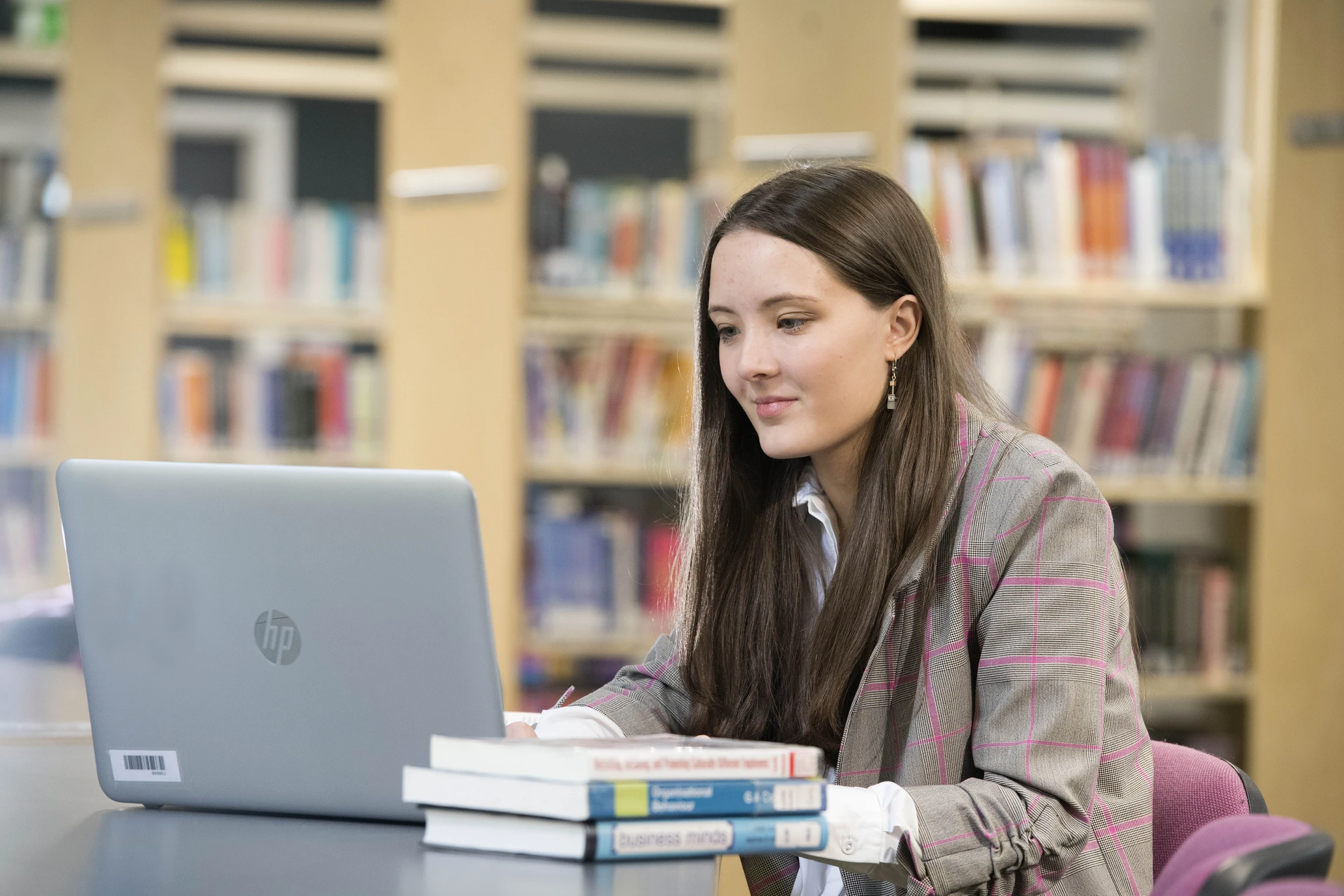 girl sat in a library at a desk with a laptop and stacked books